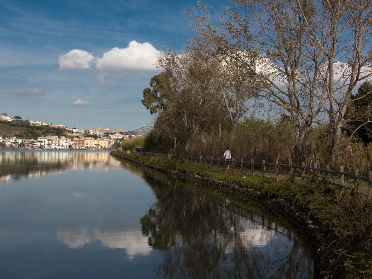 The calm waters of Lake Avernus reflecting the sky, with a lakeside path and the town of the Campi Flegrei in the distance