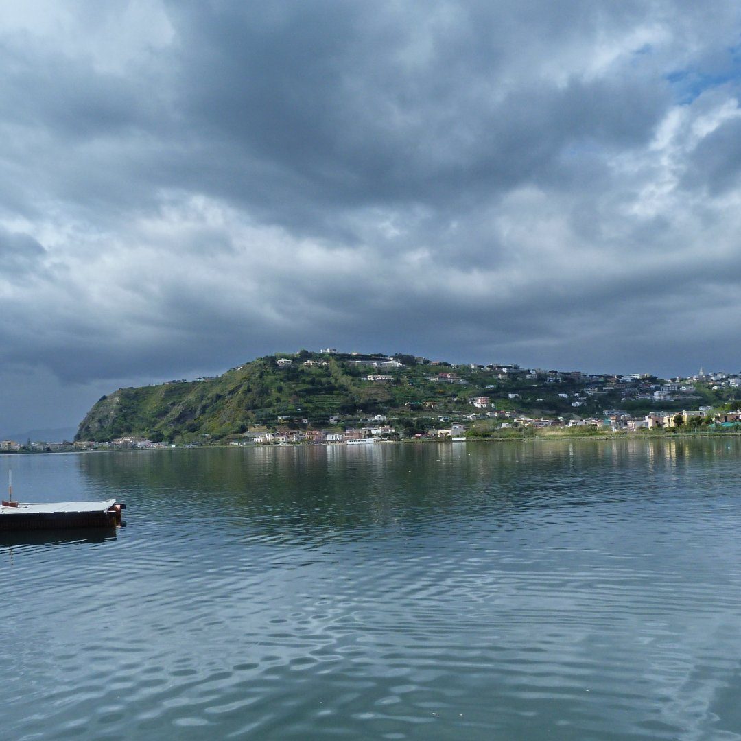 The calm green water of Lake Miseno at Bacoli under a dramatic cloudy sky, with the hillside town beyond
