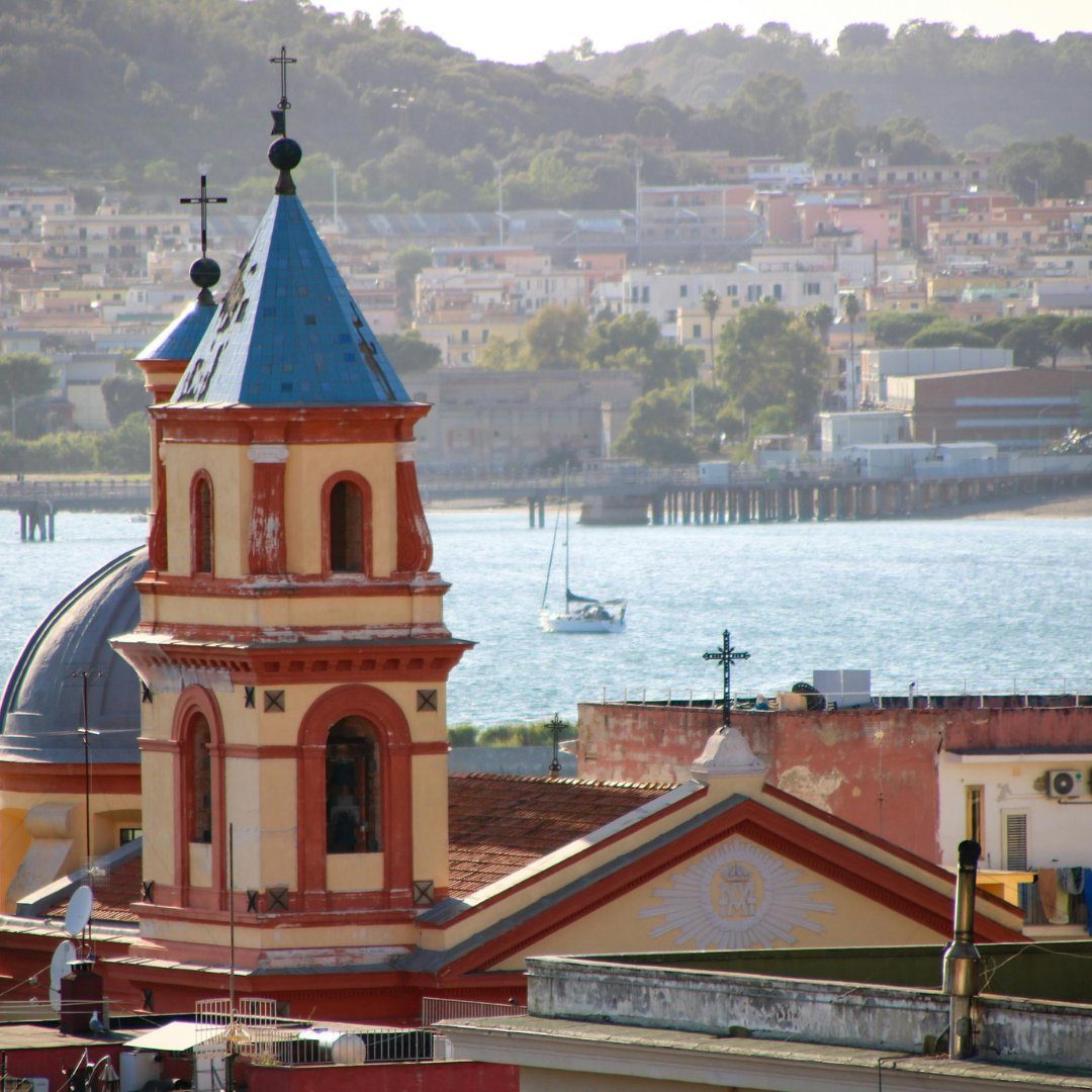 A red-and-yellow baroque bell tower with a blue majolica dome rising above the rooftops and harbor of Pozzuoli